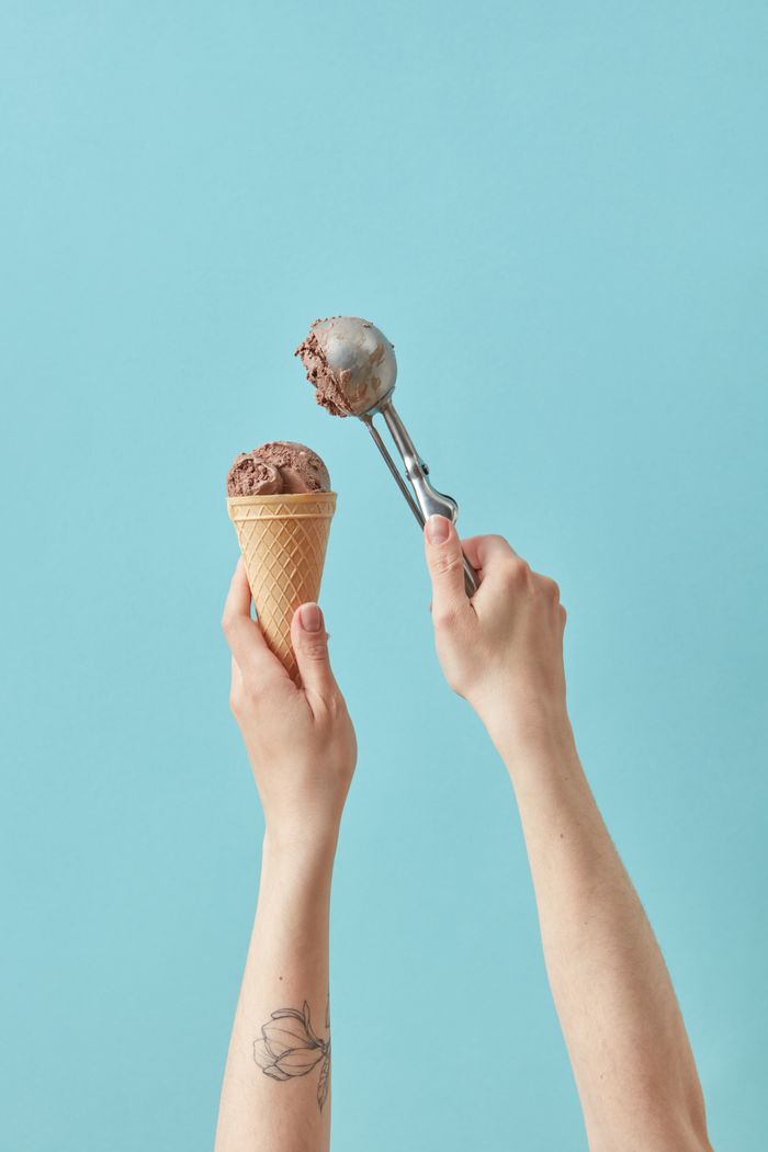 A hand holding a chocolate ice cream cone with a metal scoop above. Minimalist food photography.